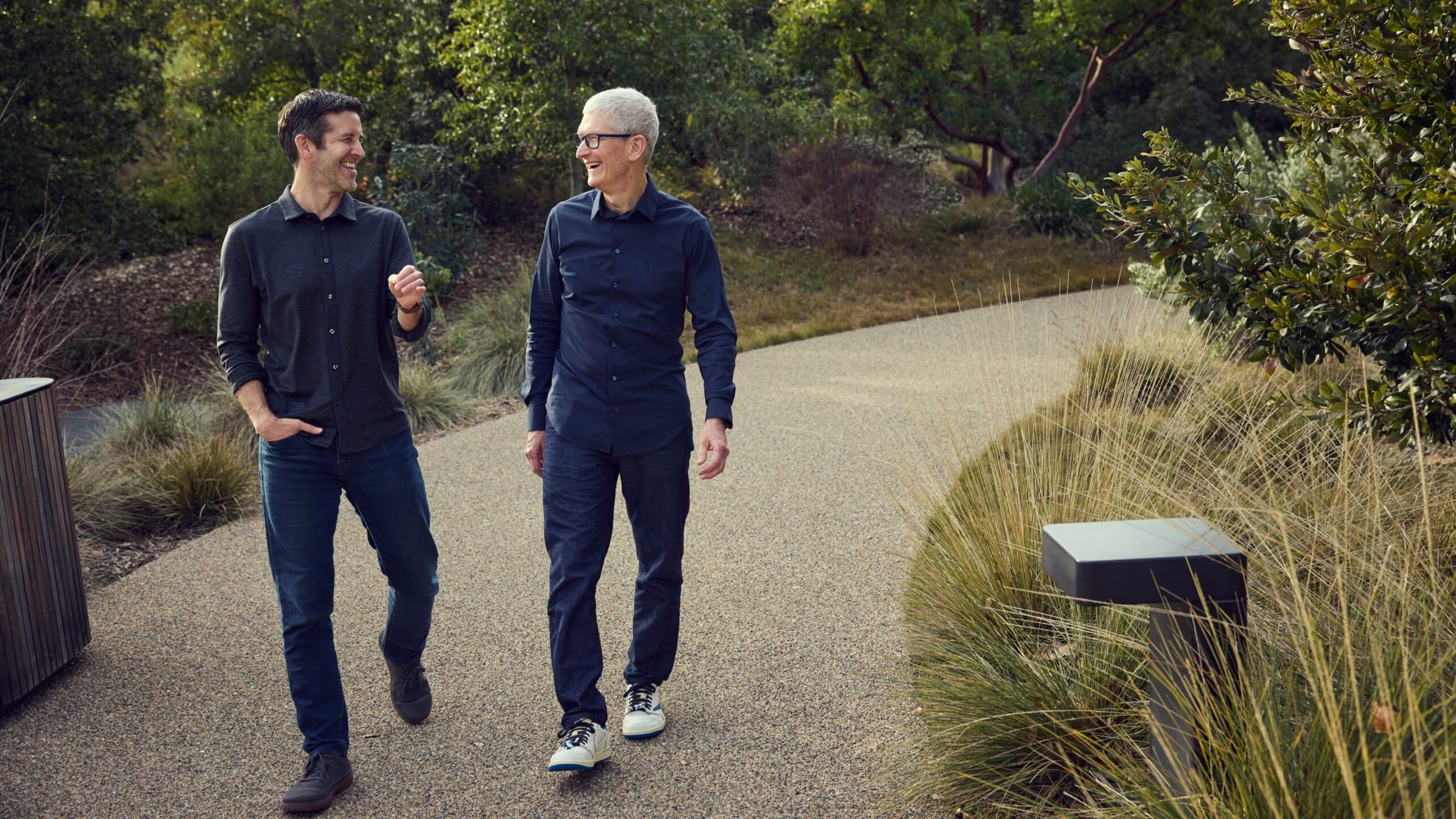 John Ternus and Tim Cook walking and laughing along a landscaped path at Apple Park