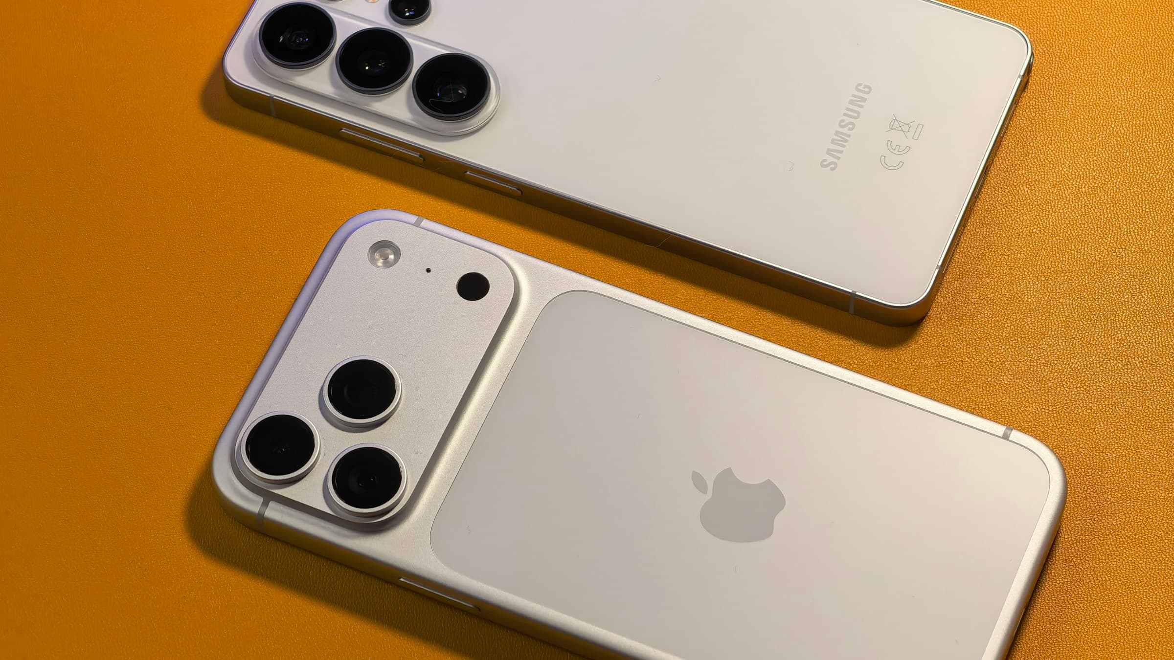 A white iPhone and a white Samsung Galaxy smartphone lying side by side on an orange surface, showing their rear cameras
