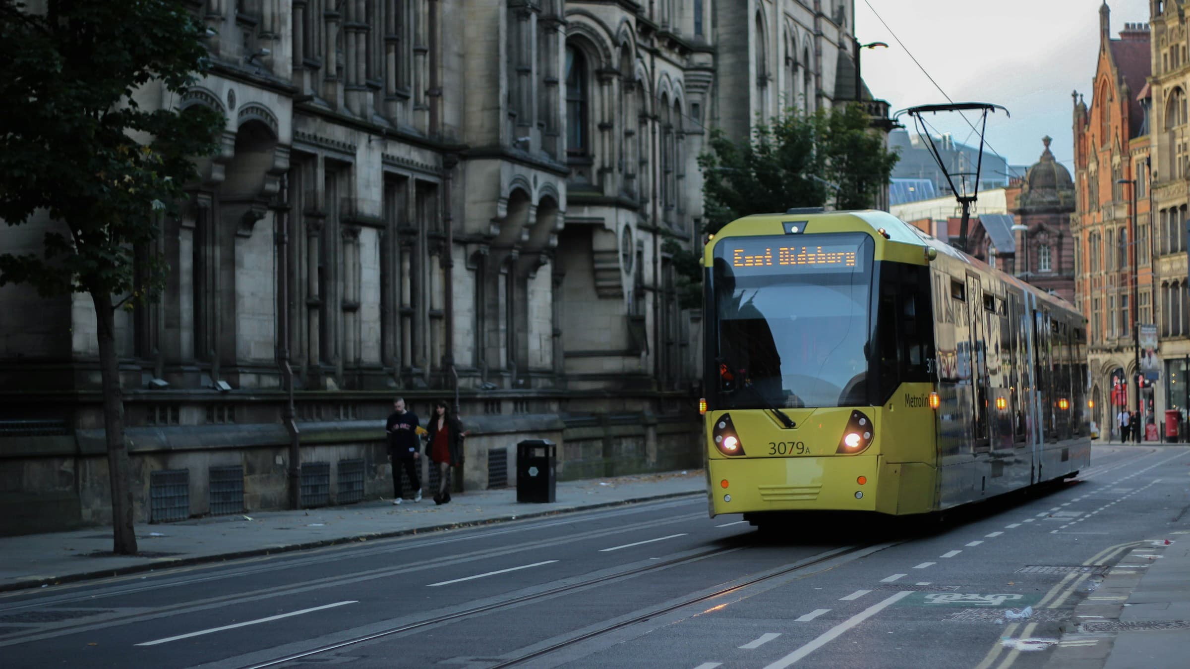 A yellow Metrolink tram passing Manchester Town Hall on a city-centre street