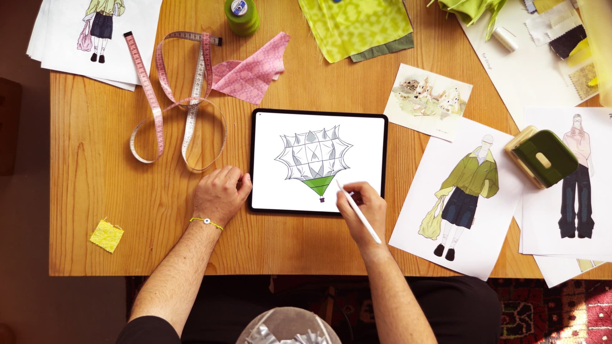 Bünyamin Aydın sketching a hot-air balloon design on iPad Pro with Apple Pencil Pro, surrounded by fashion sketches and fabric swatches