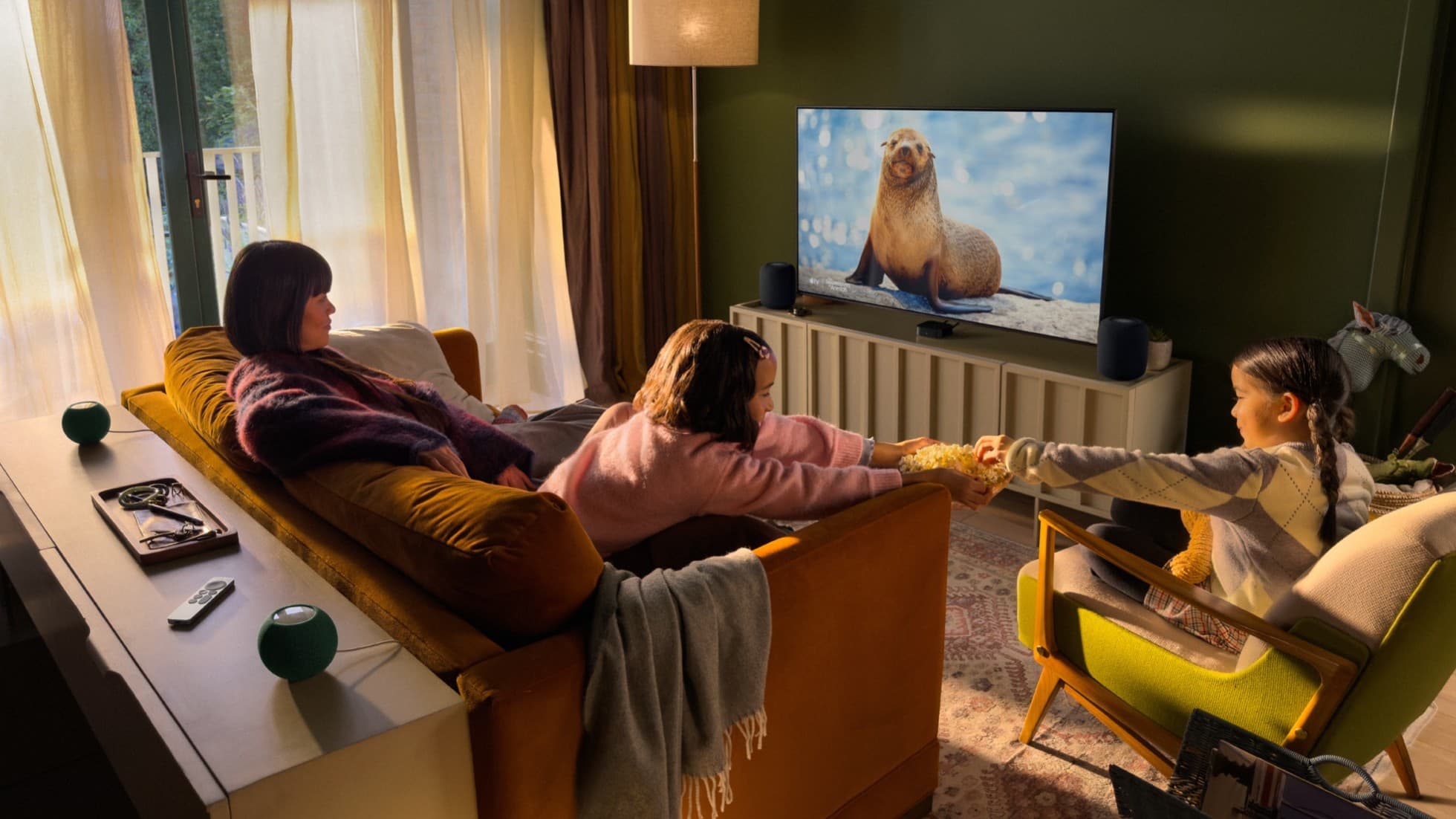 Family in a living room watching TV with the new Apple TV 4K, two HomePod speakers beside the TV and two HomePod mini speakers behind the sofa